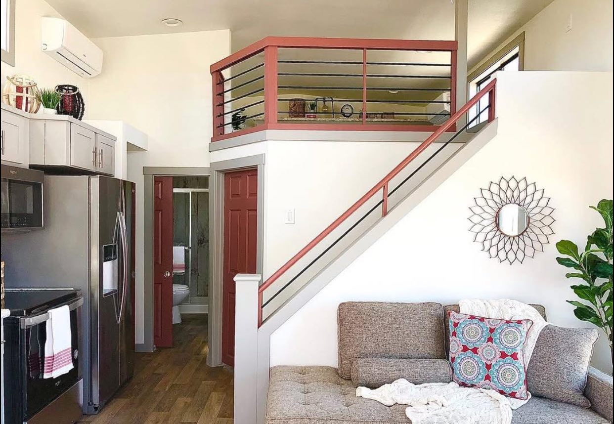 Cozy tiny home interior with a loft. Features a gray couch with colorful pillows, wall mirror, potted plant, stainless steel kitchen, and a wooden staircase.