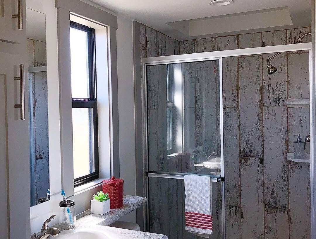 Bright, rustic bathroom with distressed wooden shower walls, sliding glass doors, and a striped towel. Decor includes a red jar and small plant.