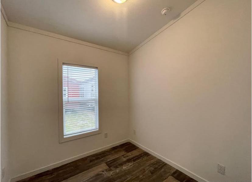 Small, empty room with beige walls and dark wood laminate flooring. A single window with blinds lets in outside light, creating a simple, tranquil ambiance.