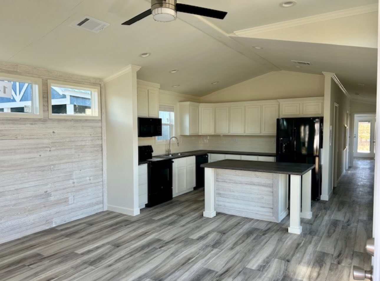 Modern kitchen with vaulted ceiling, white cabinetry, and black appliances. Light wood flooring contrasts with a textured gray accent wall. Bright, airy feel.