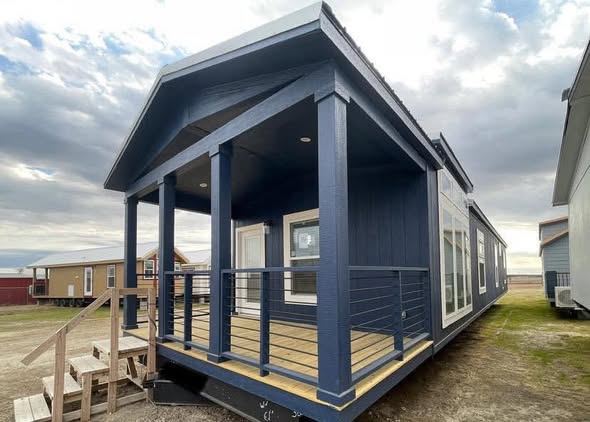 A modern, dark blue tiny house with a covered porch and railings, set against a cloudy sky. Nearby, similar homes are visible, creating a cozy scene.