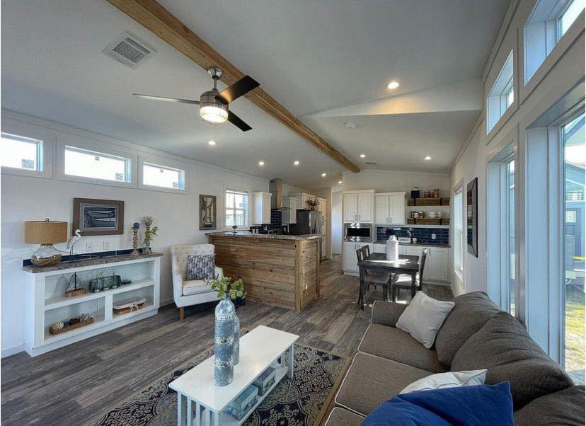 Modern open-plan living area with wood accents. Features a cozy sofa, wooden coffee table, ceiling fan, and a bright kitchen with navy backsplash. Airy, inviting ambiance.
