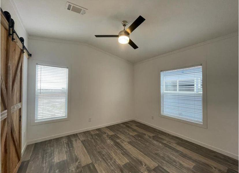 Empty room with white walls, dark wood laminate flooring, two windows with blinds, a ceiling fan with light, and a rustic wooden barn door.