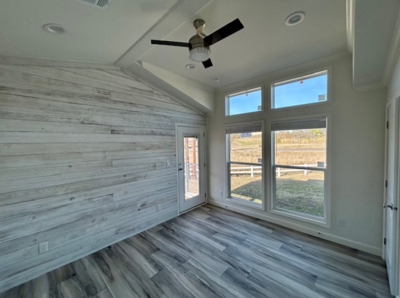 Bright room with large windows showing a grassy view, white shiplap walls, gray wood flooring, and a ceiling fan. Modern and airy atmosphere.