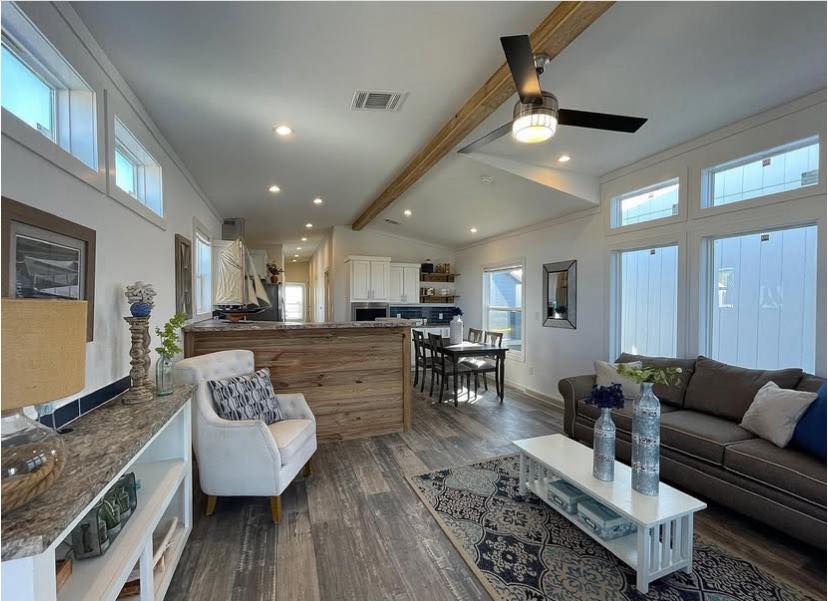 Modern living room with wood floors, a beige sofa, white coffee table, and armchair. Large windows and ceiling fan add a bright, airy feel.