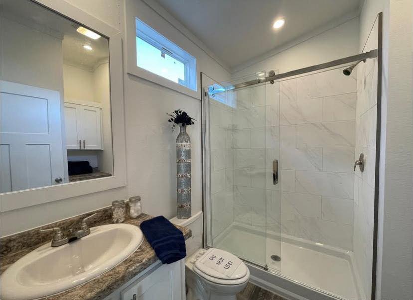 A small, modern bathroom featuring a glass-enclosed shower, a marble countertop with a sink, a tall decorative vase, and a sign on the toilet reading "Do Not Use."
