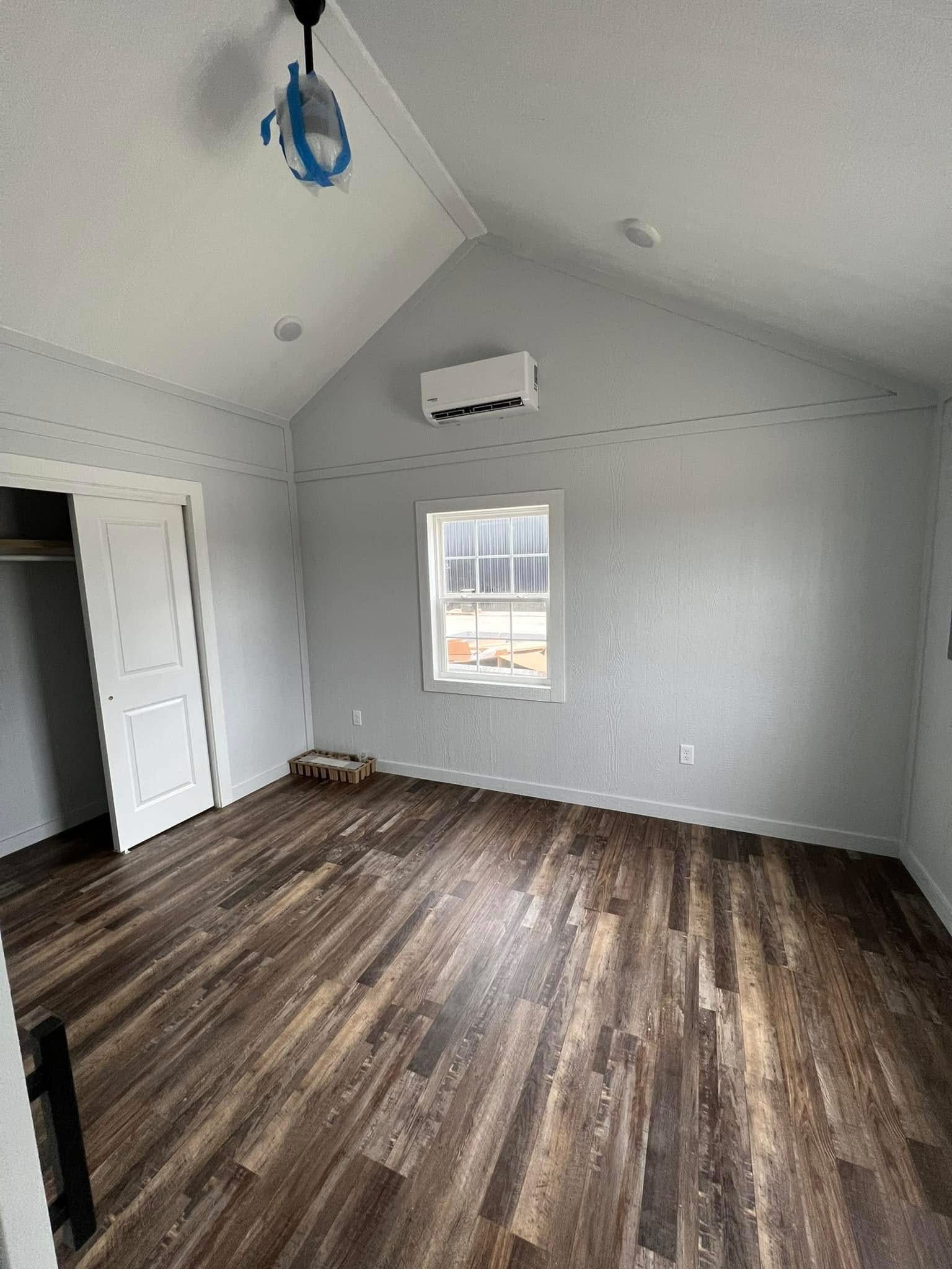 Empty room with light gray walls, wood flooring, and a single window. A closed white closet door and ceiling light fixture are visible, creating a minimalist tone.