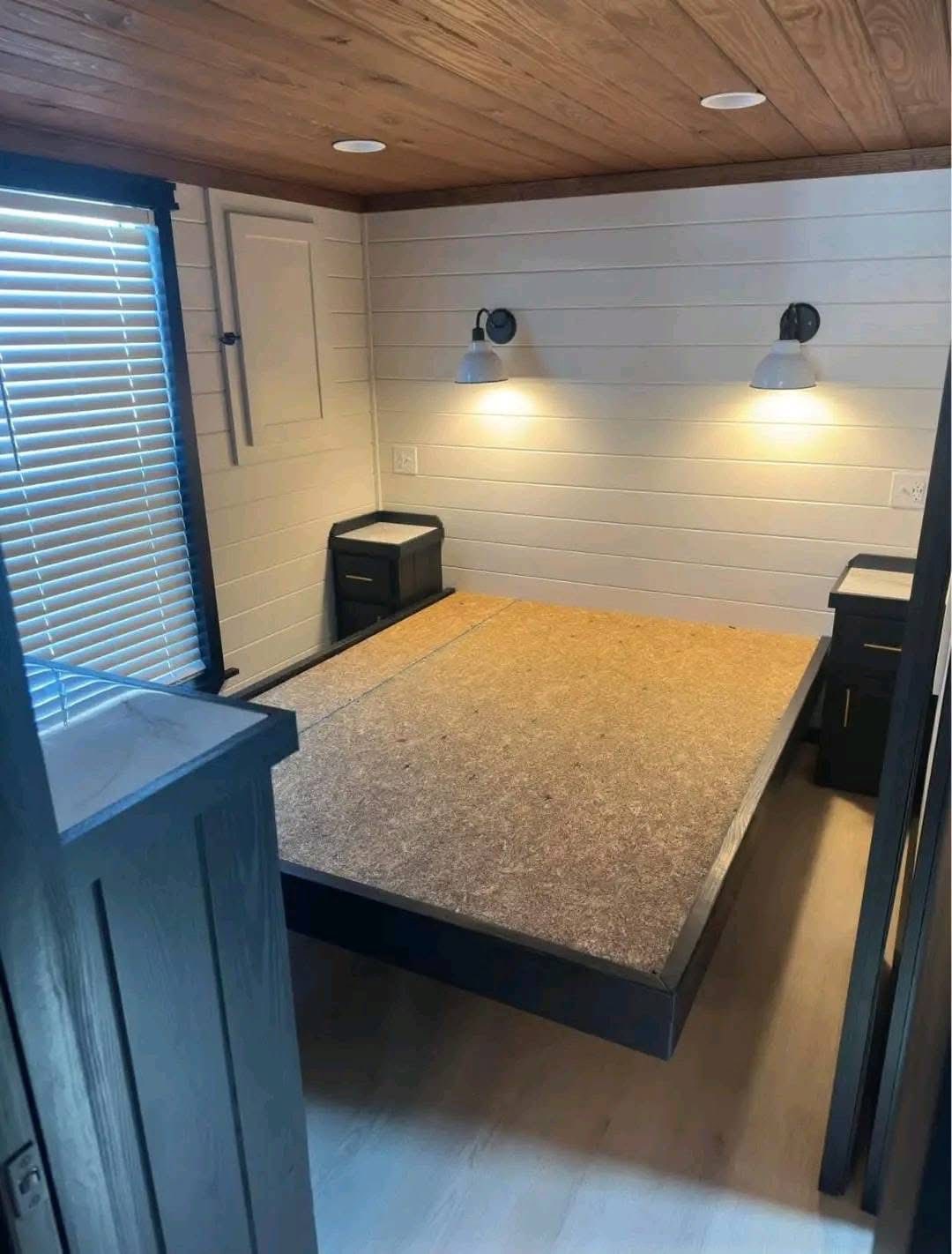 Cozy bedroom with a minimalist setup featuring a wood platform bed without a mattress, flanked by two nightstands. Soft lighting from wall-mounted lamps adds warmth, complemented by wooden walls and ceiling.