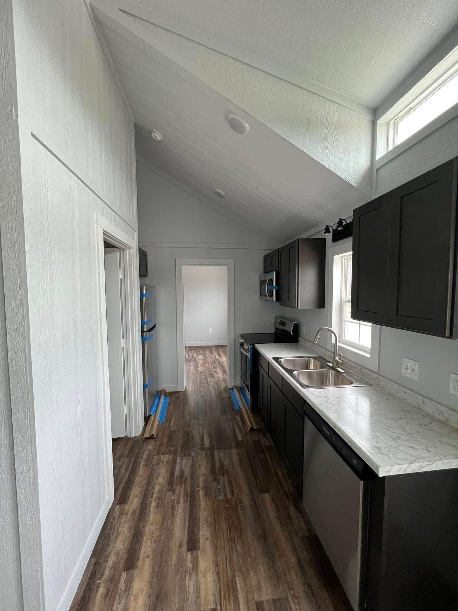 Narrow kitchen with a high slanted ceiling, featuring dark cabinets, marble countertops, and a double sink. Wooden flooring adds warmth to the modern space.