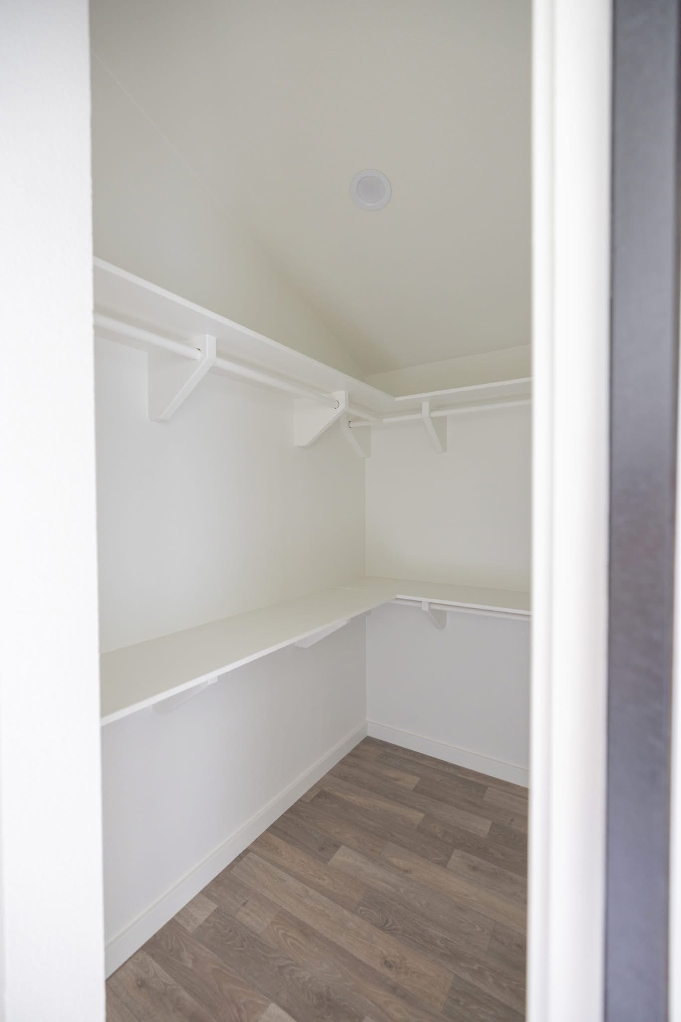 A spacious, empty walk-in closet with white walls and shelves. It features wooden flooring and a high ceiling, conveying a clean, minimalist feel.