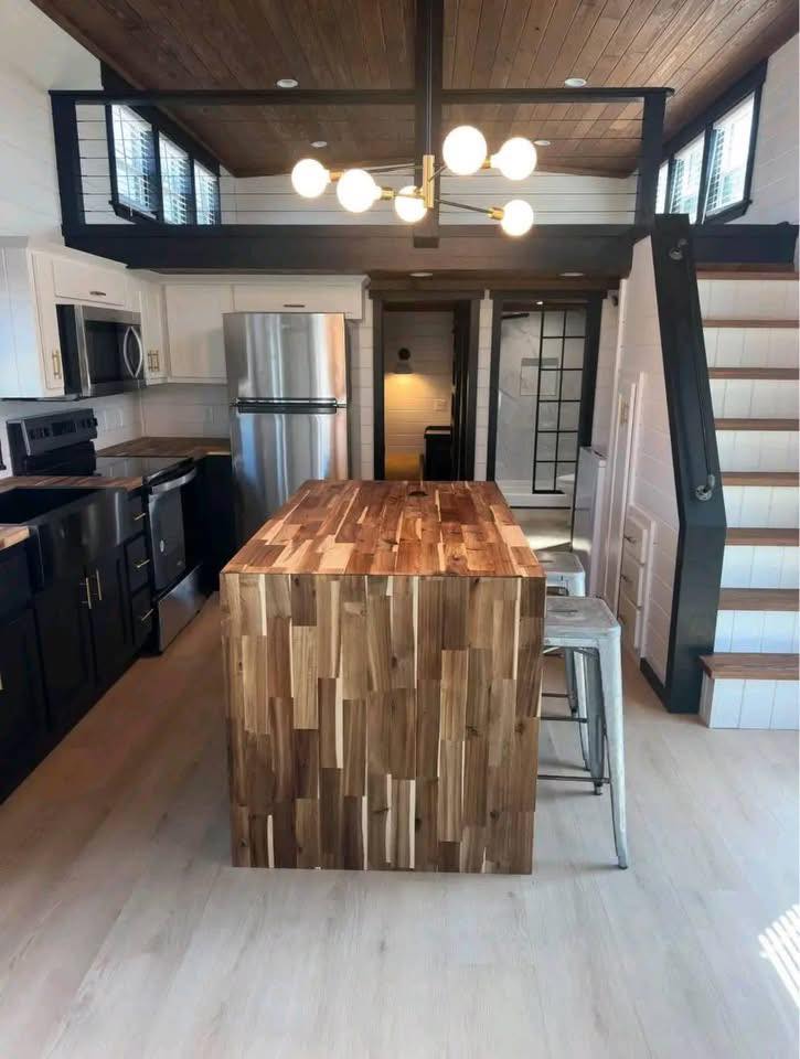 Modern kitchen with wood accents features a central wooden island, sleek appliances, a small dining area, and stairs leading to a loft. Bright, open design.
