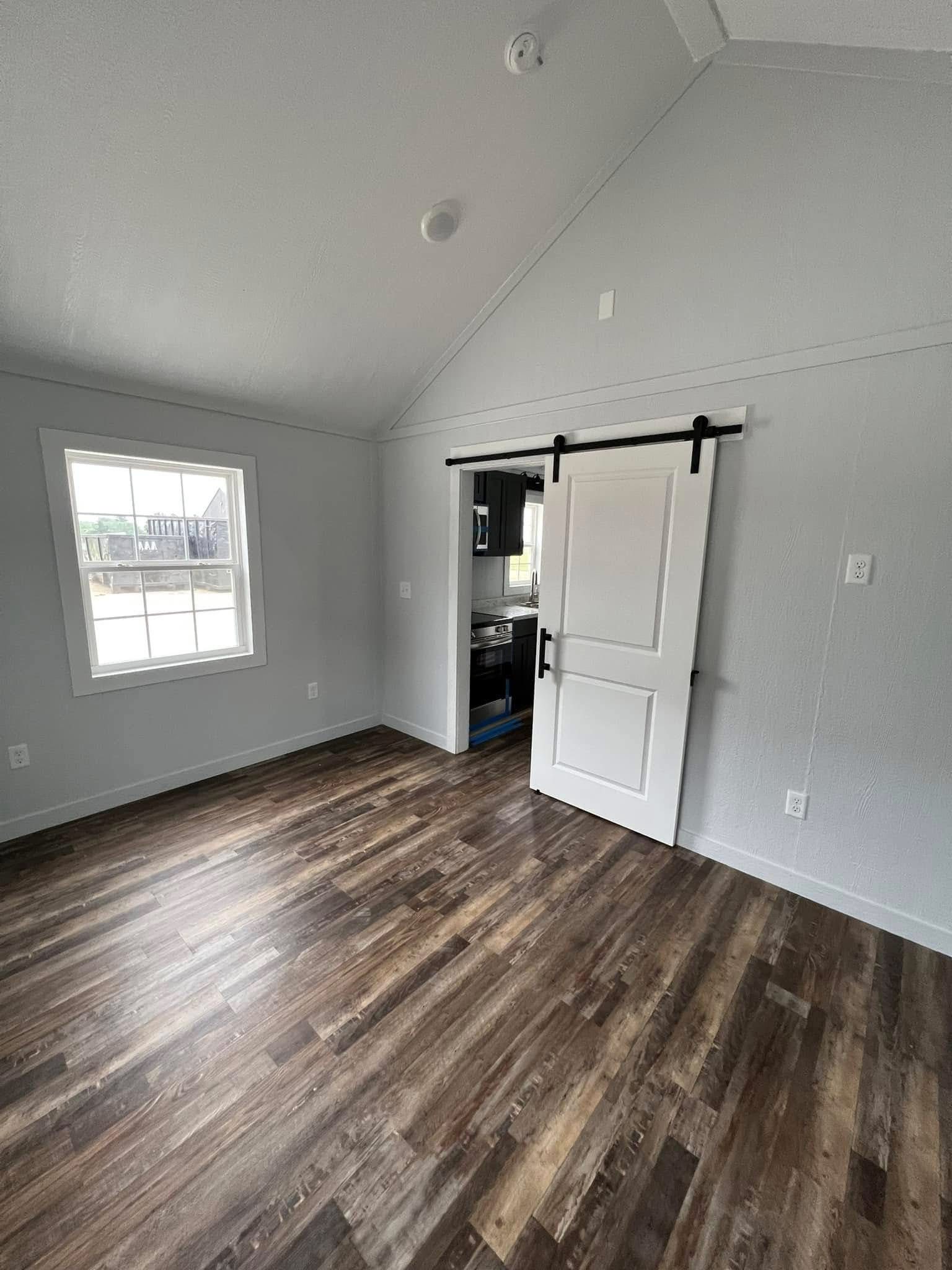 A bright, empty room with wooden floors, gray walls, and a window on the left. A white sliding barn door on the right leads to a kitchen area.