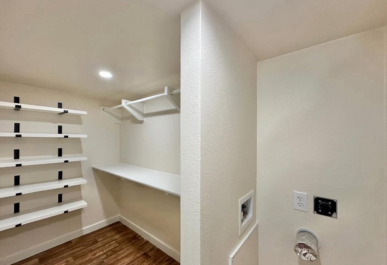 Spacious laundry room with wood floors, built-in white shelves, and a countertop. Bright lighting enhances the clean, organized feel.