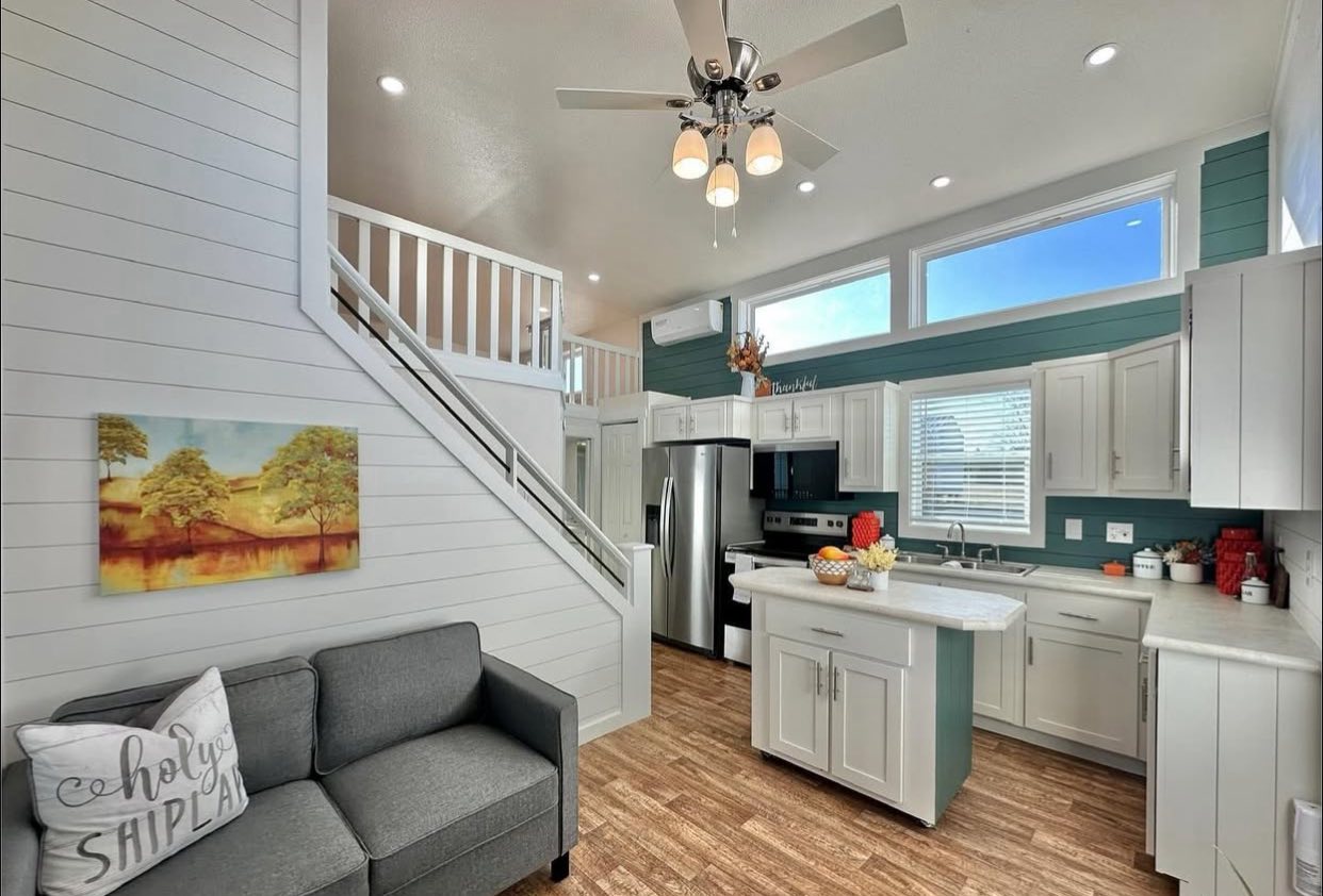 Bright modern kitchen and living area with white cabinets, teal accents, and wood flooring. Cozy gray sofa, wall art, and an inviting light fixture.