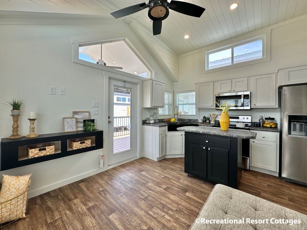 Bright, modern kitchen in tiny home with white cabinets, stainless steel appliances, and wood flooring. Black ceiling fan and natural light enhance space.