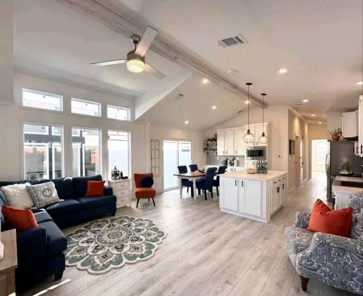 Spacious, modern open-plan living room and kitchen with light wood floors. Features a navy sofa, patterned rug, and white cabinets with pendant lights.