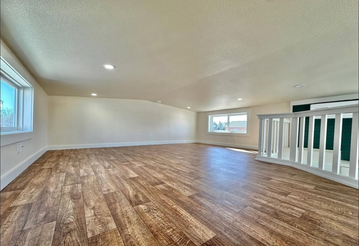Spacious loft with wood flooring, two windows providing ample natural light, low ceiling, and white railing. Bright and airy atmosphere.