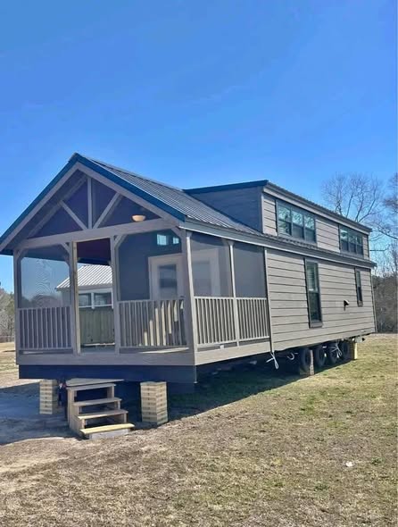Tiny house with a gabled roof on wheels, featuring a covered porch with railings. Set on a grassy field under a clear blue sky, conveying tranquility.
