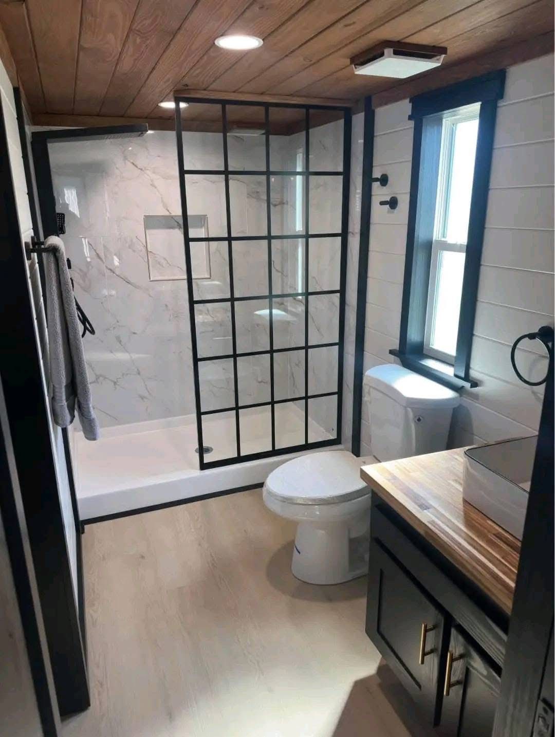 Stylish bathroom with a wood ceiling and light flooring. Features a black-framed shower, marble walls, modern fixtures, and natural light from a window.