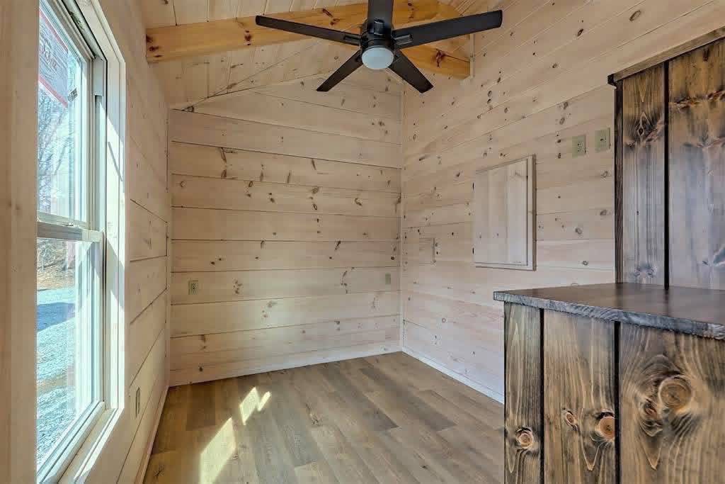 Wood-paneled room with light pine walls, a large window on the left, a black ceiling fan above, and a rustic wooden cabinet on the right.