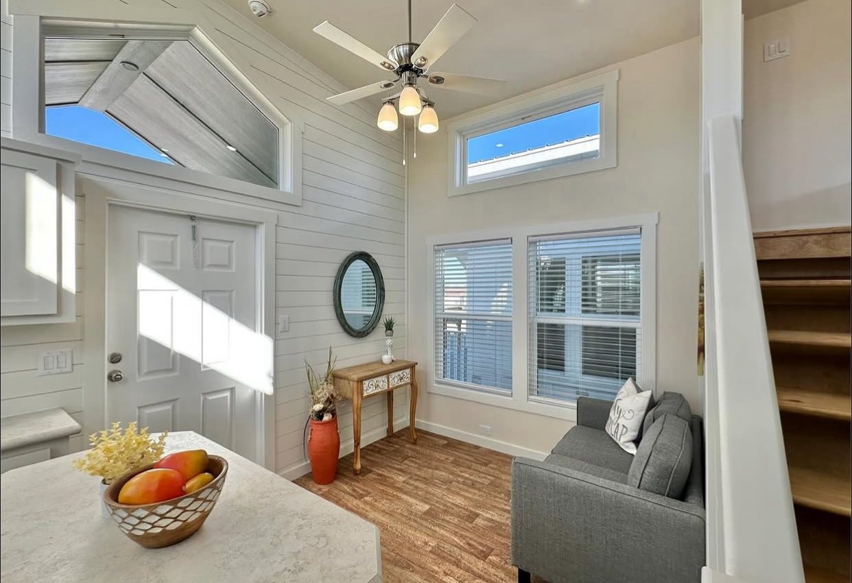 Bright tiny home interior with wood floors, high ceilings, and large windows. Features a gray sofa, decorative table with a mirror, and a ceiling fan. Cozy and inviting atmosphere.