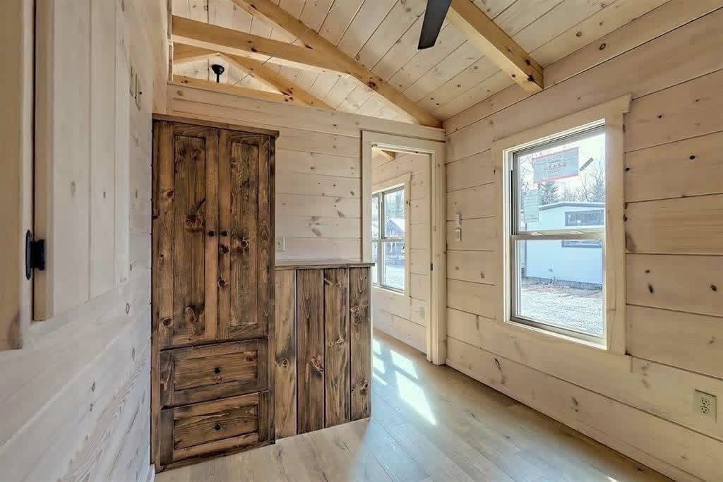 Cozy wooden interior of a small cabin with natural light streaming through two windows. Features a rustic wooden cabinet and vaulted ceilings.