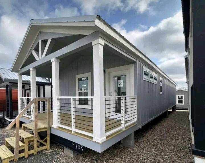 A modern tiny house on a cloudy day, featuring a gray exterior, white trim, and a small porch with railings. It rests on a gravel lot, conveying simplicity.