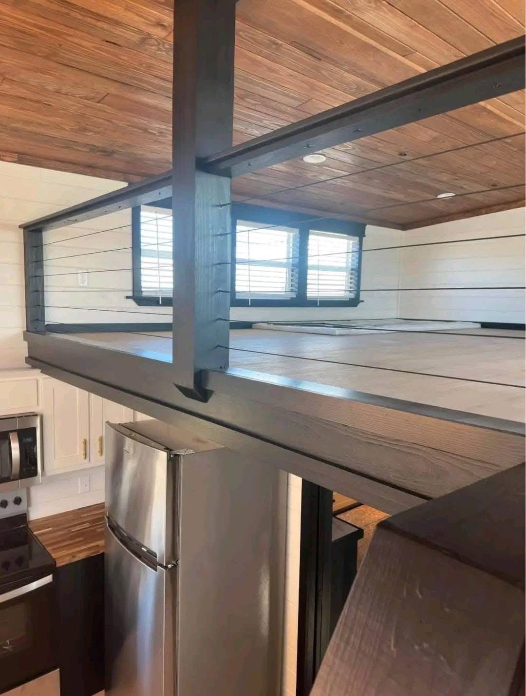 Loft interior with a wooden ceiling and floor, featuring a sleek metal railing. A kitchen with a stainless steel fridge is visible below. Bright and cozy.