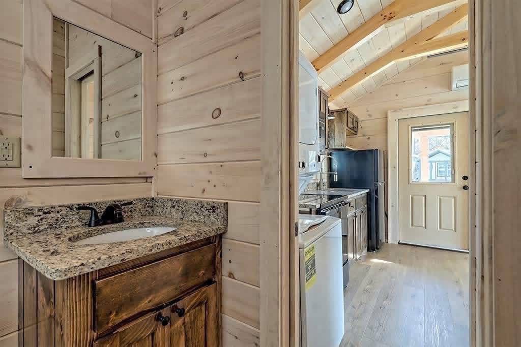 Cozy cabin interior with wooden walls and ceiling. A small bathroom with a granite sink is visible, leading into a narrow kitchen with light streaming through a door.