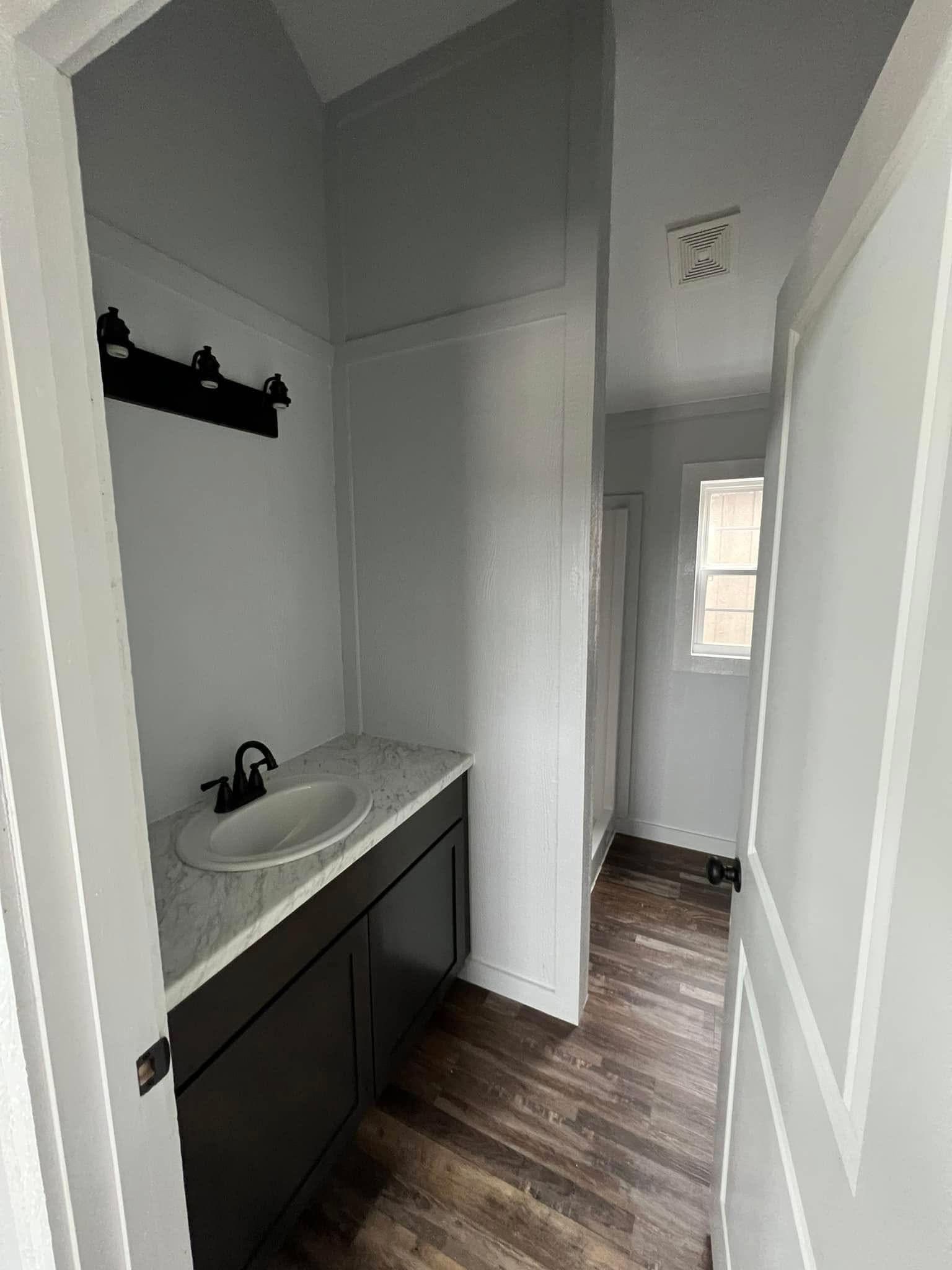 Narrow bathroom with a dark vanity and white marble countertop on the left. Light gray walls, dark wood floor, and a small window on the right. Simple, modern design.