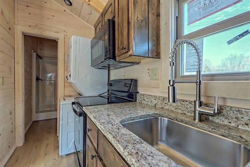 Compact kitchen in a tiny home featuring light wood walls, a granite counter, stainless steel sink, stacked washer-dryer, oven, and microwave.
