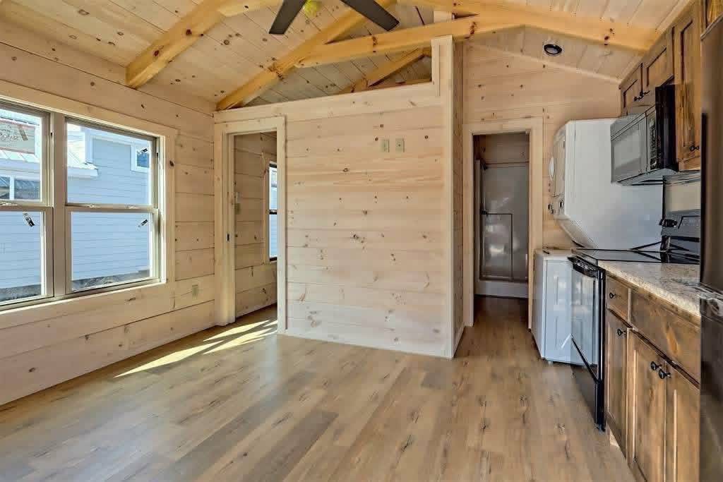 Cozy wooden interior of a small cabin with light wood walls, vaulted ceiling, and laminate floors. Kitchen area features dark cabinets and appliances.