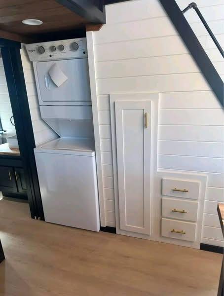 Compact laundry area in a white-paneled room features a stacked washer and dryer beside sleek storage cabinets with gold handles, conveying efficient use of space.