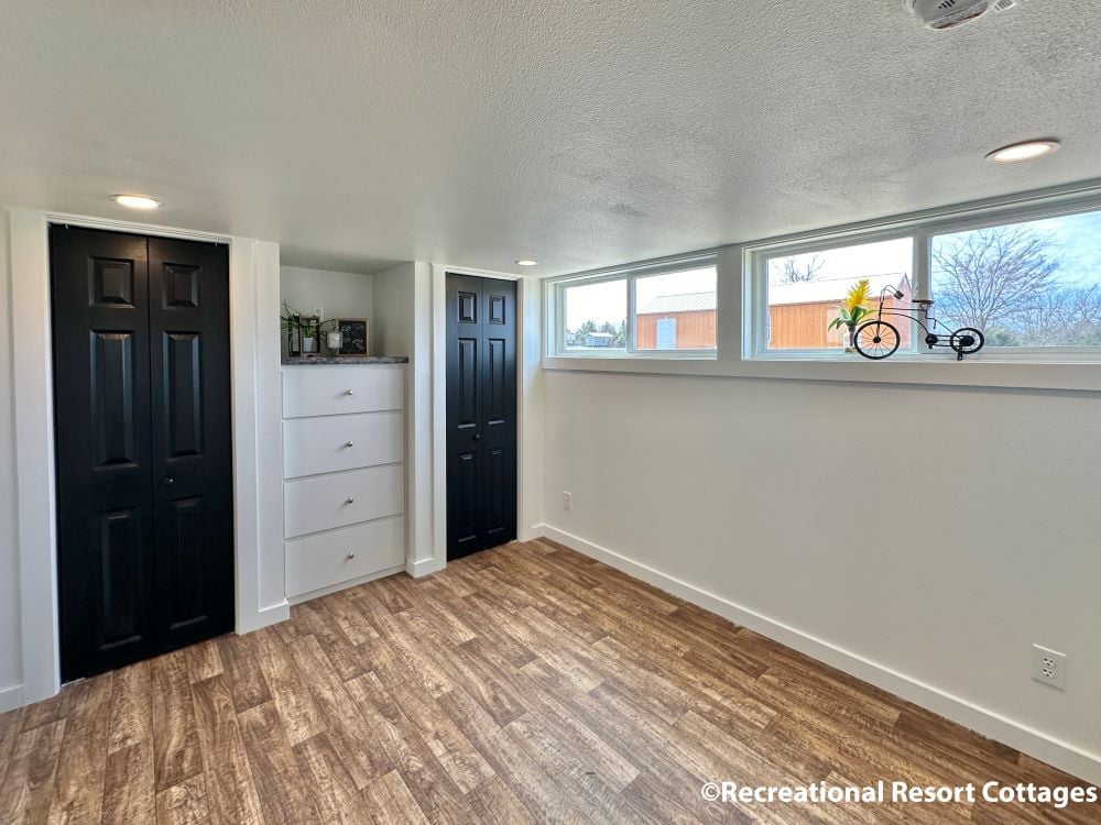 Bright room with wood flooring, black closet doors, and white walls. High windows allow natural light. Decorative bike and flowers on the sill. Cozy and modern.