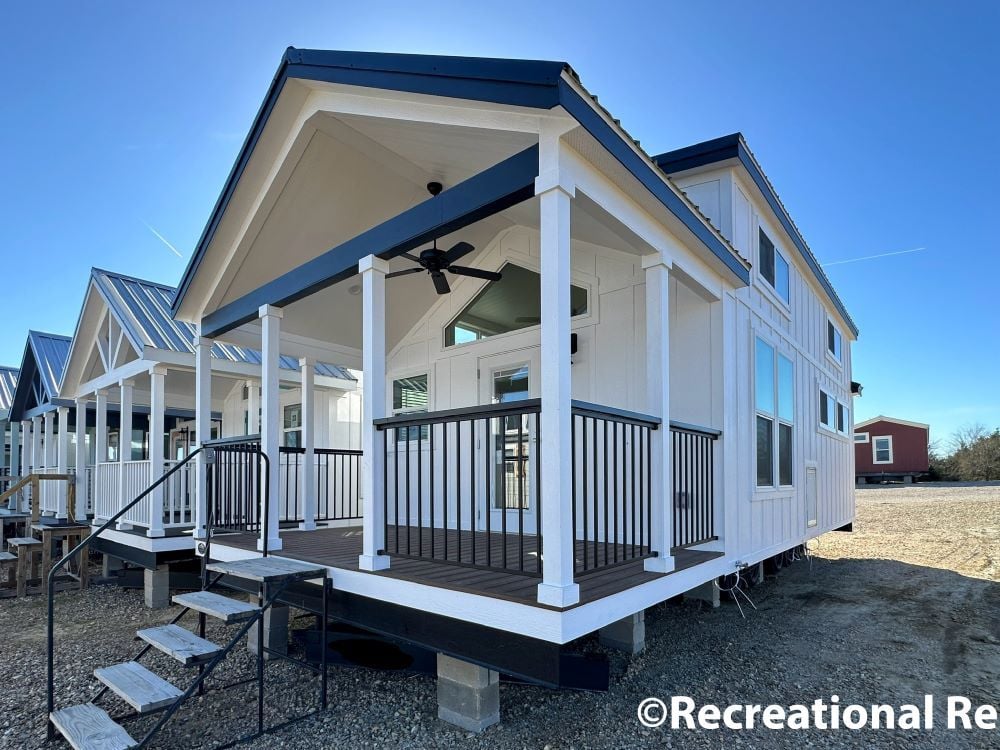 A row of modern tiny homes with white siding and dark roofs under a clear blue sky. Prominent front porch with railings and steps, evoking a cozy, inviting feel.