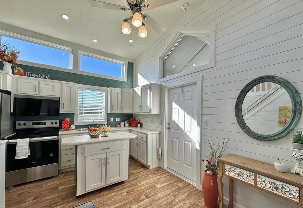 Bright kitchen with white cabinets, wood floors, and high windows. A round mirror hangs on shiplap walls above a console table with decor. Cozy and inviting.