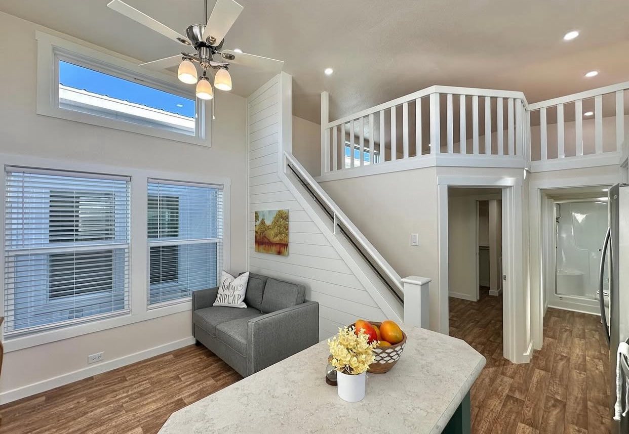 A cozy, modern tiny home interior featuring a loft with white railing, stairs, a gray couch, large windows, ceiling fan, and a kitchen island with fruit.