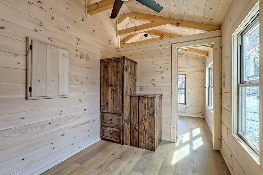 Rustic wooden room interior with light wood paneling and a small window letting in natural light. A dark wood cabinet stands against the wall.