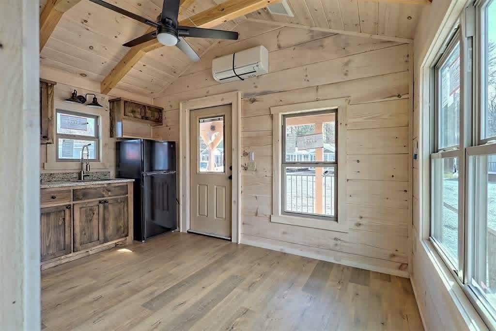 Rustic tiny house interior with natural wood walls and ceiling. A compact kitchen features a small fridge and sink. Light floods through windows, creating a cozy and inviting atmosphere.