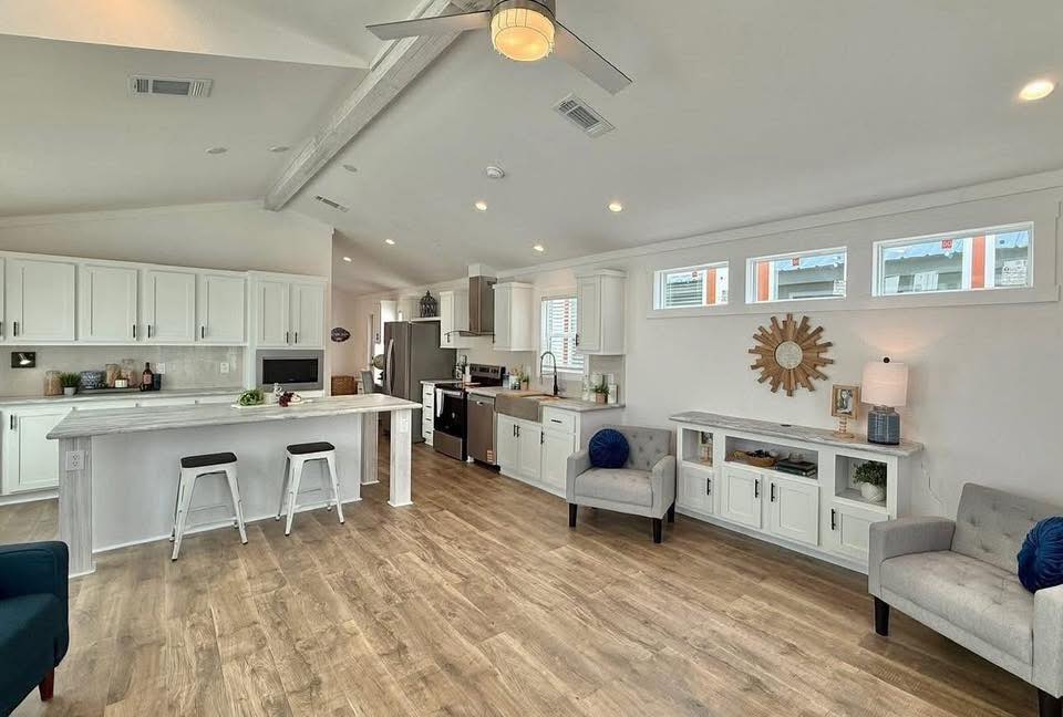 Spacious modern kitchen and living area with white cabinets, a large island with stools, light wood flooring, and cozy armchairs. Bright and inviting.