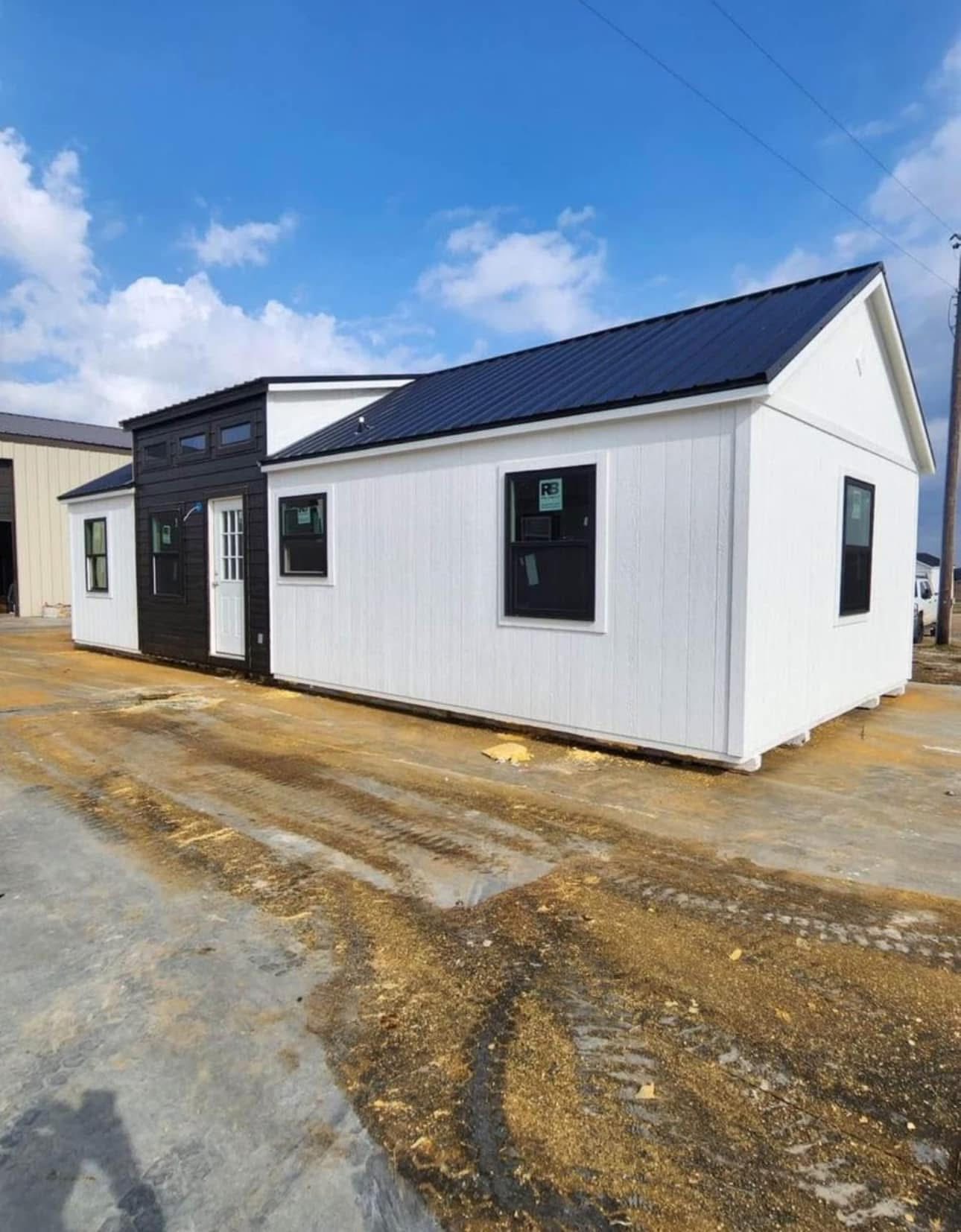A modern, single-story tiny house with white siding and a black door stands on a concrete lot under a blue sky with scattered clouds.