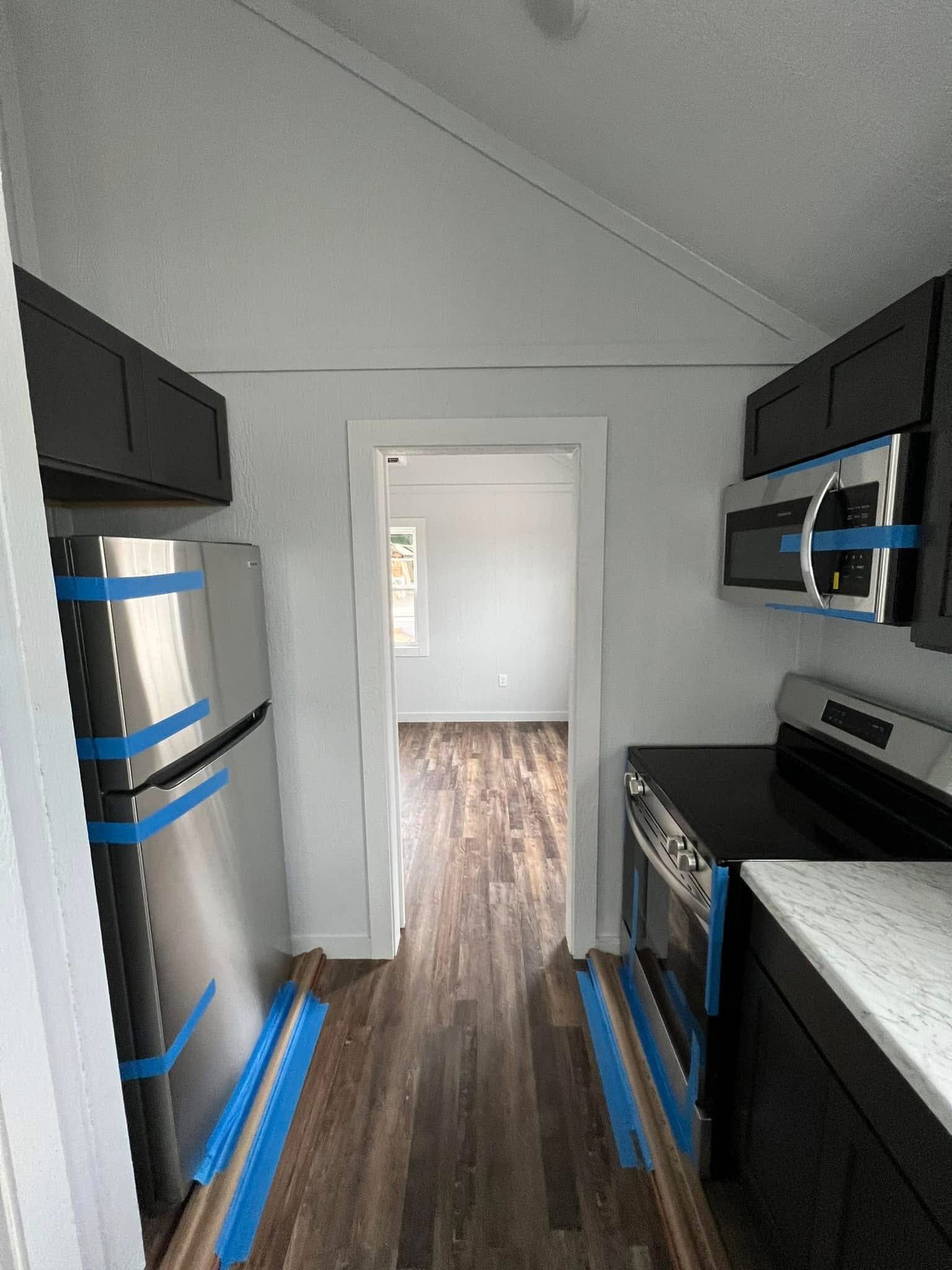 Narrow kitchen with dark cabinets and stainless steel appliances, marked with blue tape, and wood flooring. Bright adjacent room visible.