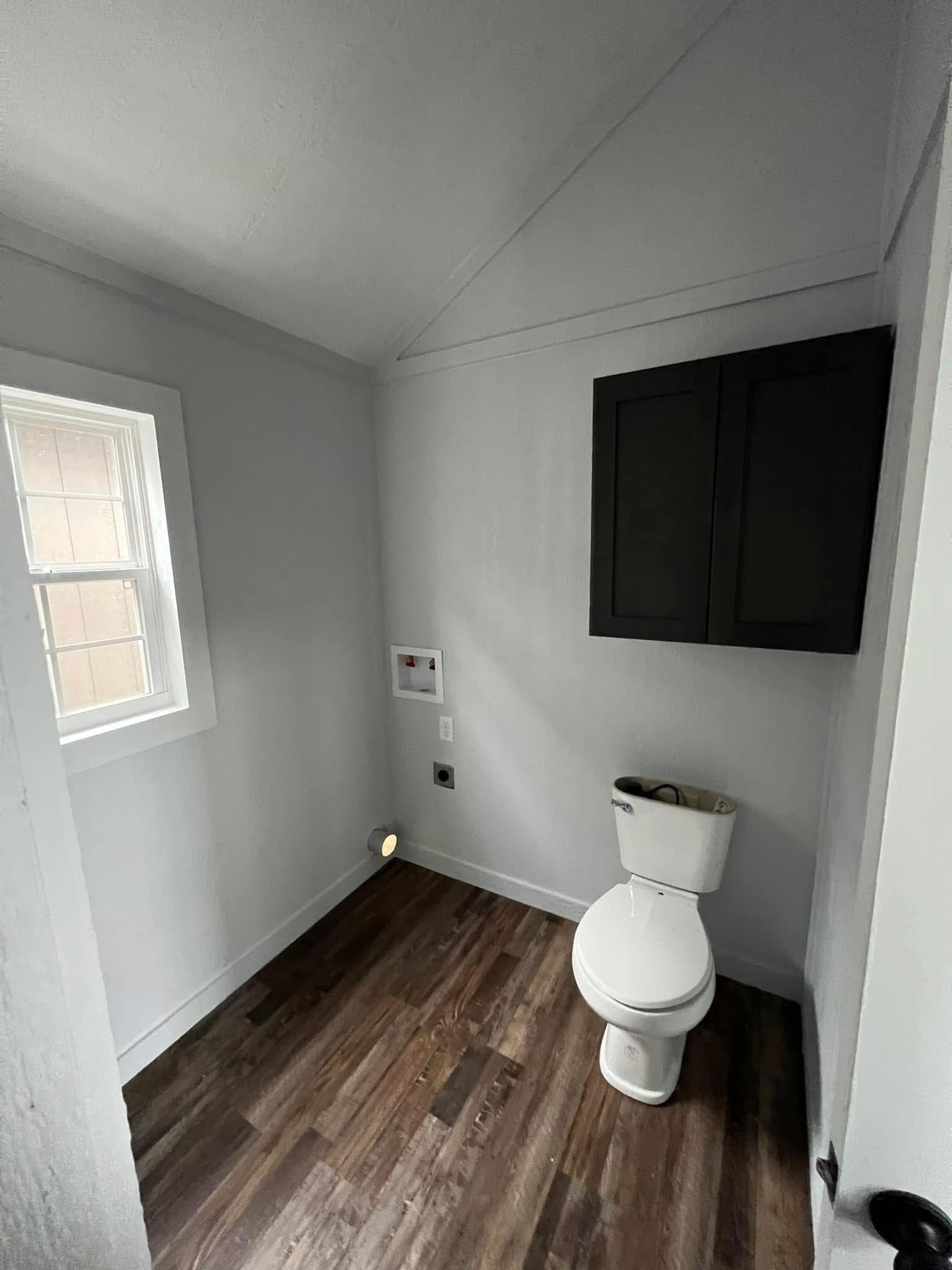 A small bathroom with a white toilet on right, dark wooden cabinet above, and a small window on the left. The room has light gray walls and wood flooring.