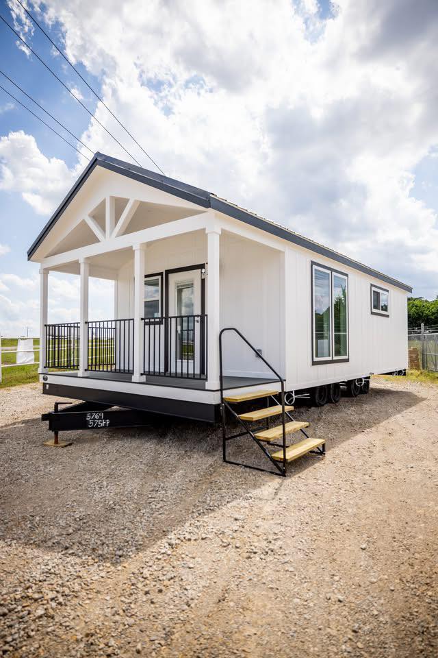 White tiny house on wheels with a gray roof, black railing, and steps leading to a small porch. It's set on a gravel path under a partly cloudy sky.