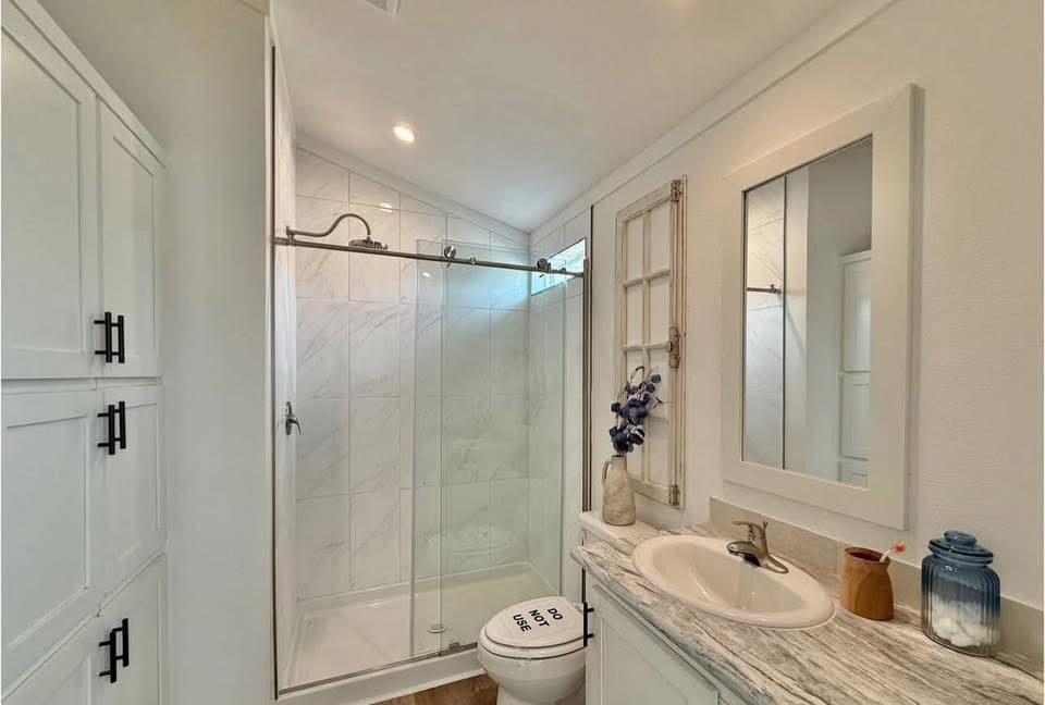 Compact modern bathroom with marble countertop, white cabinet, glass shower, and decorative items. Toilet seat cover says "Do Not Use."