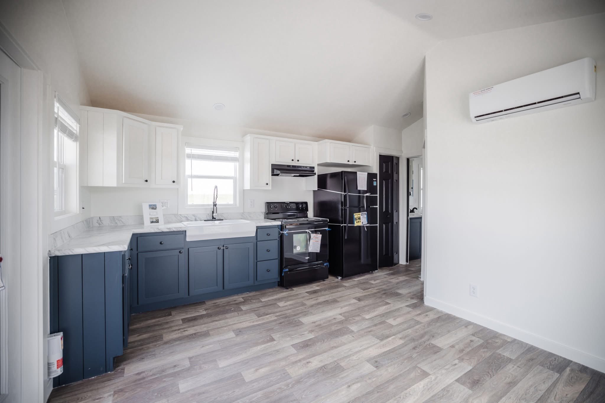 Modern kitchen with blue and white cabinets, a farmhouse sink, and black appliances on wooden floors. Bright light from a window enhances the airy feel.