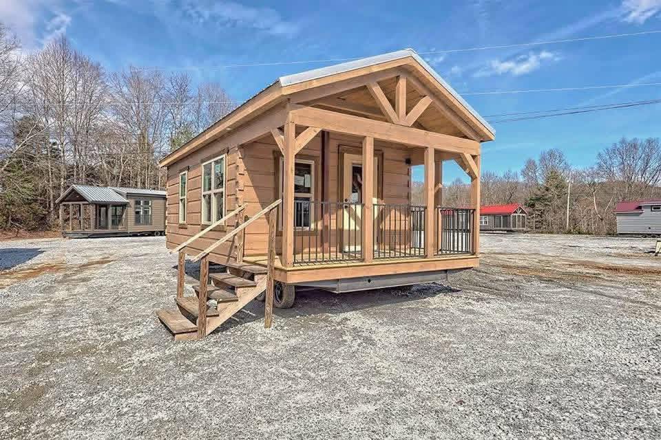 A small, wooden cabin with a porch and railing sits in a gravel lot under a clear blue sky. Other cabins and leafless trees are visible in the background.