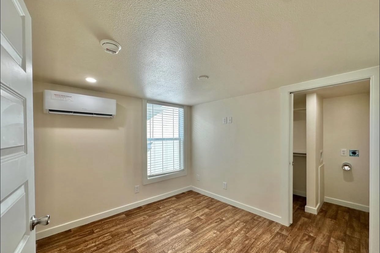 Small, empty room with light wood flooring and beige walls. A window with blinds allows natural light. An air conditioning unit is mounted on the left wall.