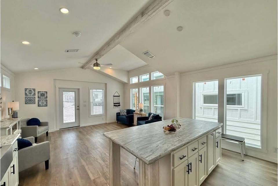 Bright, spacious living room with light wood flooring, cozy gray and blue seating, art on walls, large windows, and a kitchen island with marble top.