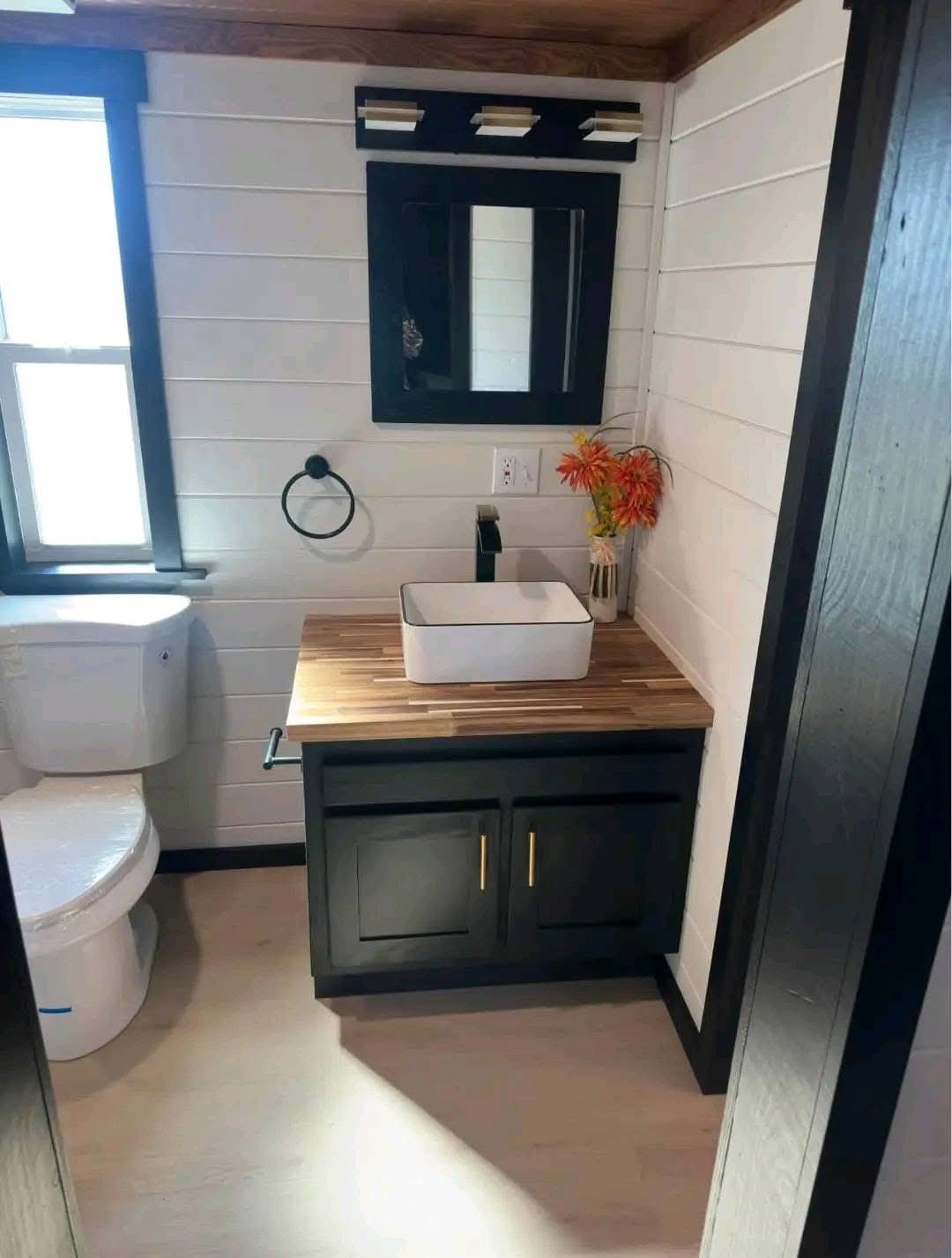 Compact bathroom with a white toilet, wooden vanity, square sink, and black cabinet. A mirror and lights hang above, with orange flowers for decor.
